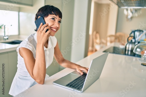 Beautiful brunette woman with short hair at home using computer laptop speaking on the phone