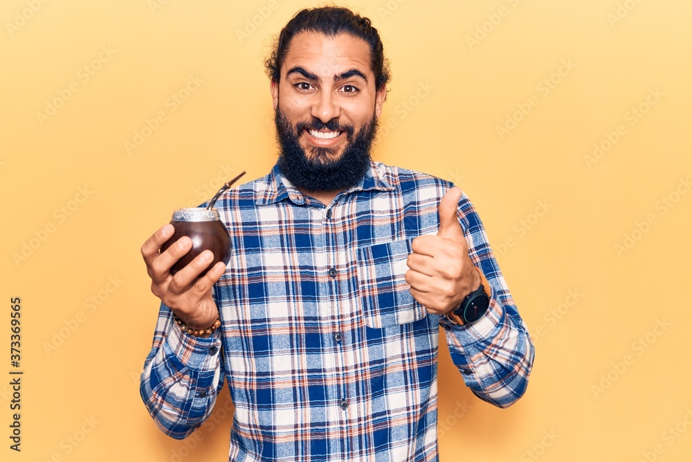 Young arab man drinking mate infusion smiling happy and positive, thumb up doing excellent and approval sign