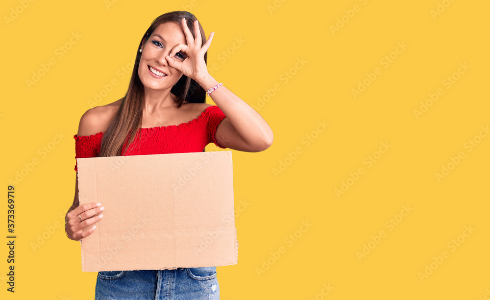 Young beautiful hispanic woman holding banner cardboard smiling happy doing ok sign with hand on eye looking through fingers
