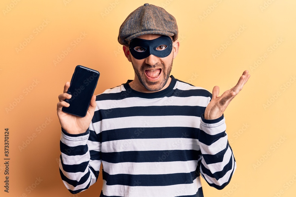 Young handsome bald man wearing burglar mask holding smartphone celebrating achievement with happy smile and winner expression with raised hand Young handsome bald man wearing burglar mask holding smartphone celebrating achievement with happy smile and winner expression with raised hand