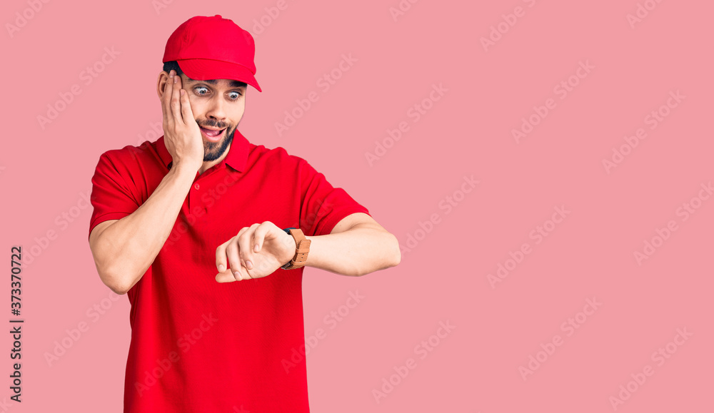 Young handsome man with beard wearing delivery uniform looking at the watch time worried, afraid of getting late