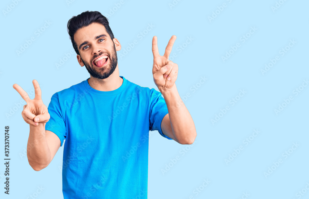 Young handsome man with beard wearing casual t-shirt smiling with tongue out showing fingers of both hands doing victory sign. number two. Young handsome man with beard wearing casual t-shirt smiling with tongue out showing fingers of both hands doing victory sign. number two.