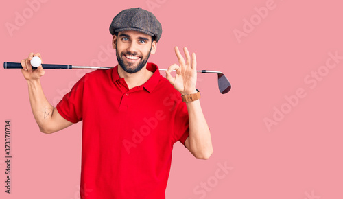 Young handsome man with beard playing golf holding club and ball doing ok sign with fingers, smiling friendly gesturing excellent symbol