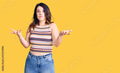 Beautiful young brunette woman wearing casual clothes clueless and confused expression with arms and hands raised. doubt concept.