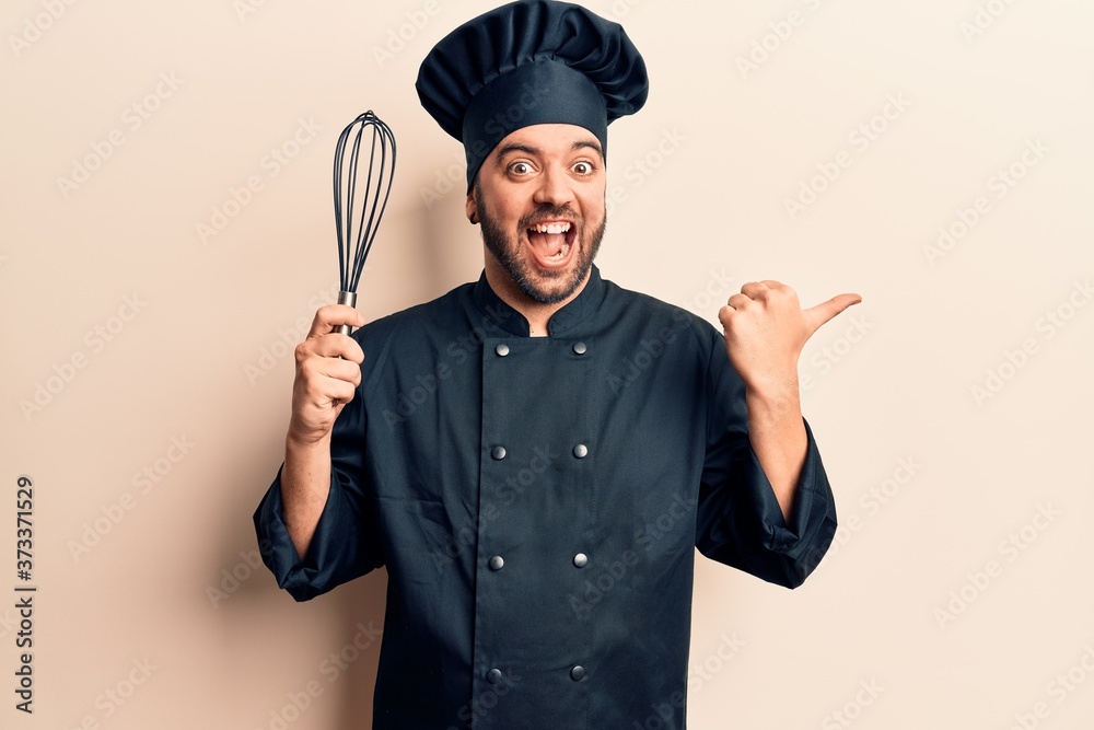 Fototapeta premium Young hispanic man wearing cooker uniform holding whisk pointing thumb up to the side smiling happy with open mouth