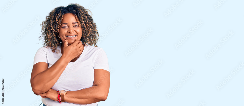 Naklejka premium Young african american plus size woman wearing casual clothes looking confident at the camera smiling with crossed arms and hand raised on chin. thinking positive.