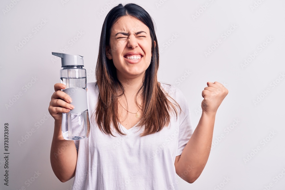 Young beautiful brunette woman drinking bottle of water over isolated white background screaming proud, celebrating victory and success very excited with raised arm