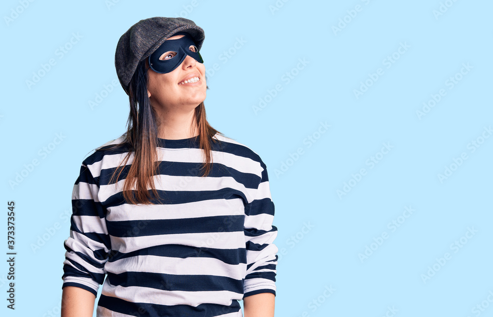 Young beautiful brunette woman wearing burglar mask looking away to side with smile on face, natural expression. laughing confident. Young beautiful brunette woman wearing burglar mask looking away to side with smile on face, natural expression. laughing confident.
