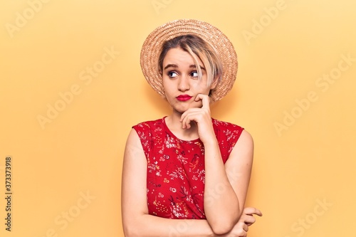 Young beautiful blonde woman wearing summer hat serious face thinking about question with hand on chin, thoughtful about confusing idea