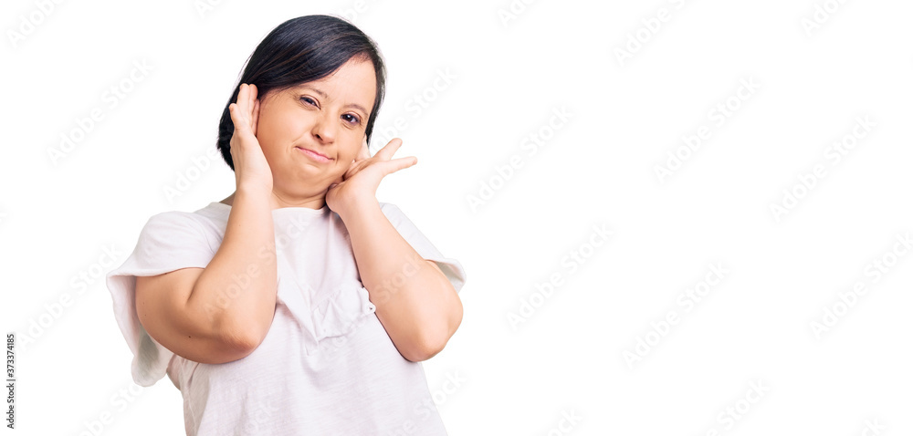 Brunette woman with down syndrome wearing casual white tshirt covering ears with fingers with annoyed expression for the noise of loud music. deaf concept. Brunette woman with down syndrome wearing casual white tshirt covering ears with fingers with annoyed expression for the noise of loud music. deaf concept.