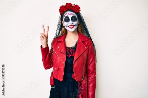 Woman wearing day of the dead costume over white smiling looking to the camera showing fingers doing victory sign. number two.