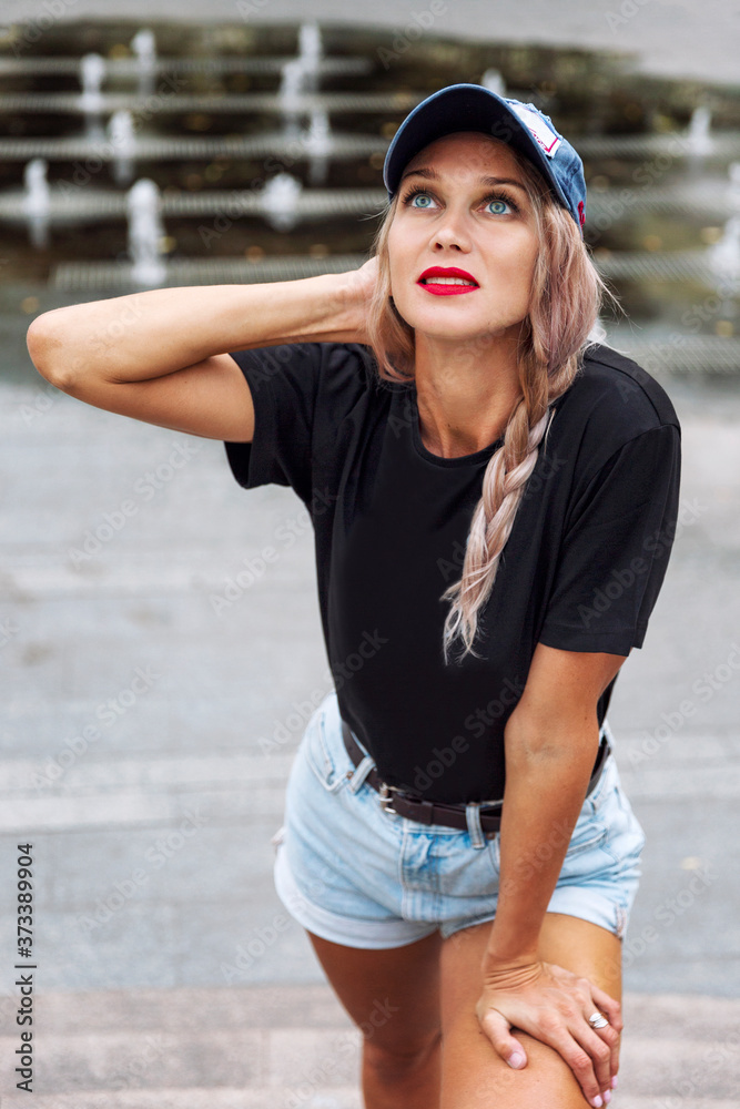 Young blonde with a braid in shorts and a cap. Beautiful smiling woman on the street of the summer city. Vertical.