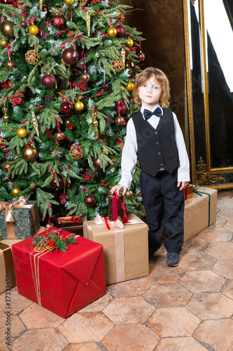 boy wearing in black bowtie, whire shirt, black suit and shoes stands near decorated xmas tree photo