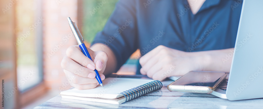 Woman is writing on a notebook with a pen in the office.On the table there are mobile phones and laptop computers.