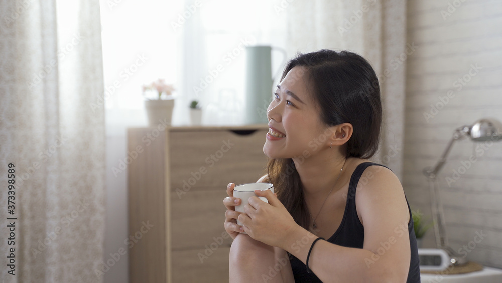 asian young lady smiling with satisfaction is having coffee in leisure. portrairt beautiful woman curling up and placing mug on knees while looking afar and thinking. asian young lady smiling with satisfaction is having coffee in leisure. portrairt beautiful woman curling up and placing mug on knees while looking afar and thinking.