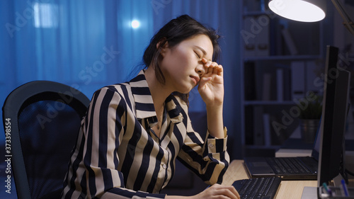 asian businesswoman is opening eyes wide trying to concentrate while working overtime at midnight. taiwanese lady propping head is rubbing sore eyes while using desktop. healthcare and work from home photo