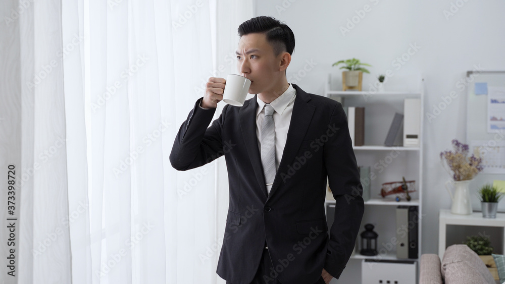 male ceo is sipping morning coffee by window with serious expression. asian man in black suit with hand in pocket is looking outside in contemplation male ceo is sipping morning coffee by window with serious expression. asian man in black suit with hand in pocket is looking outside in contemplation