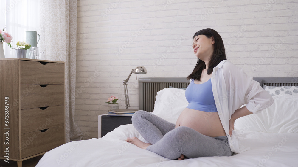 asian expectant mom is rubbing her back and groaning with painful expression in the bedroom. health problems and pregnancy concept asian expectant mom is rubbing her back and groaning with painful expression in the bedroom. health problems and pregnancy concept