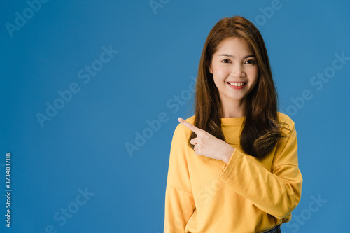 Portrait of young Asian lady smiling with cheerful expression, shows something amazing at blank space in casual clothing and looking at camera isolated over blue background. Facial expression concept.