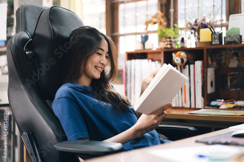 Happy smile young adult freelancer asian woman reading a book on relax in workplace at home.