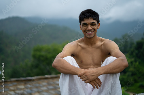 young shredded man sitting wearing a dhoti and glasses with blurred mountains in the background.