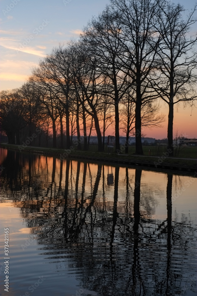 Panorama of a gorgeous scenic sunset at a river or canal, with gold and blue color in the sky and the line of trees reflected in the water. High quality photo