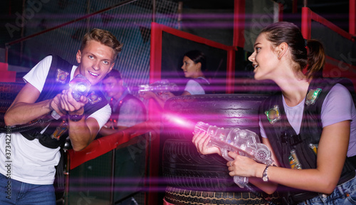 Smiling diligent positive young friends playing laser tag game with colored laser guns near tires in labyrinth