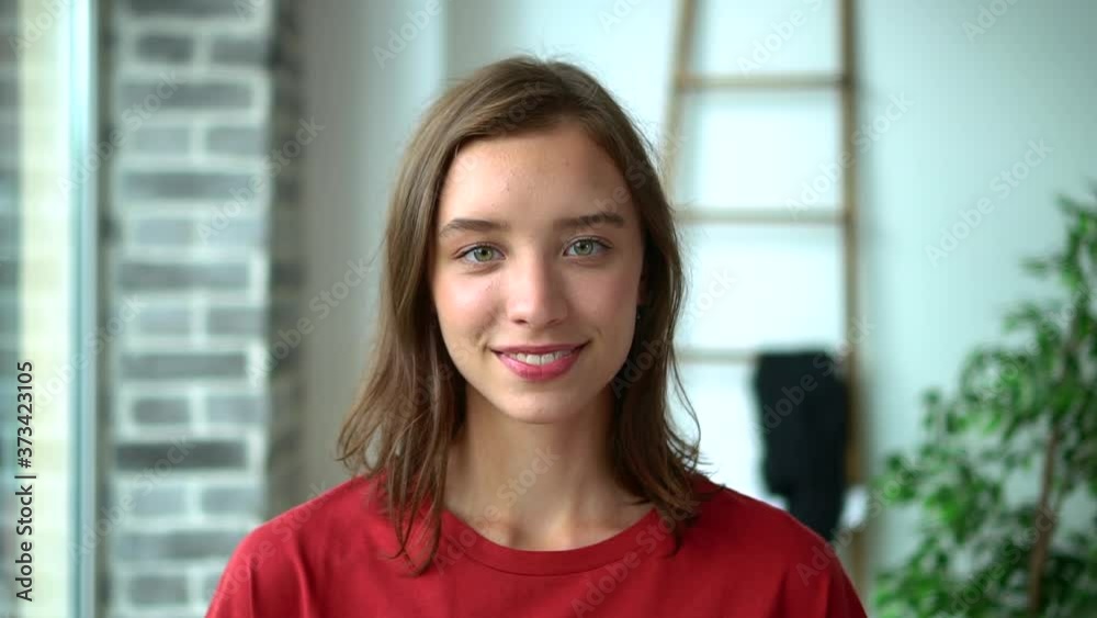 Portrait of beautiful young woman posing in front of camera while standing in light interior avki. Face view of American girl looks forward with toothy smile and poses at room with daylight. One Portrait of beautiful young woman posing in front of camera while standing in light interior avki. Face view of American girl looks forward with toothy smile and poses at room with daylight. One