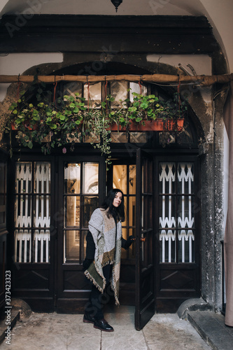 A young caucasian girl in an autumn dark coat and a wide scarf with black gloves stands in the doorway and looks into the distance. Old door with glass. Lviv, Italian courtyard, Ukraine.