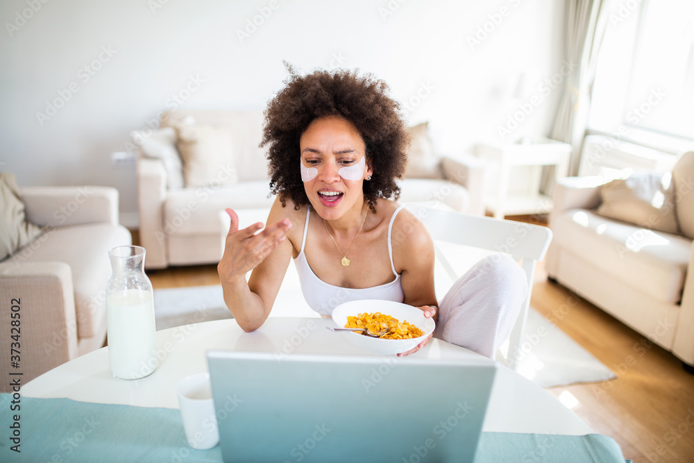 Image of happy young amazing woman sitting indoors at the table with laptop holding corn flakes. Looking at laptop computer and talking to her friends via video call. Image of happy young amazing woman sitting indoors at the table with laptop holding corn flakes. Looking at laptop computer and talking to her friends via video call.