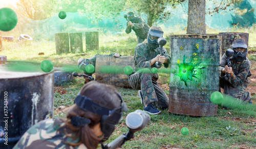Team of happy cheerful smiling adult people playing paintball on battlefield outdoor, running with guns