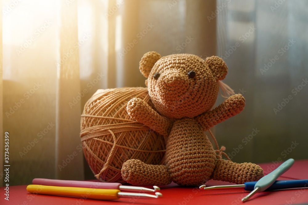 Handcrafted cute crochet brown bear doll using polyester yarn, amigurumi knitted bear doll with crochet hooks on red table - obrazy, fototapety, plakaty 