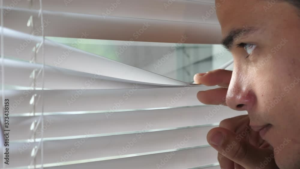 Young man looking out the window through the blinds to the street, spying. suspected or watching over.