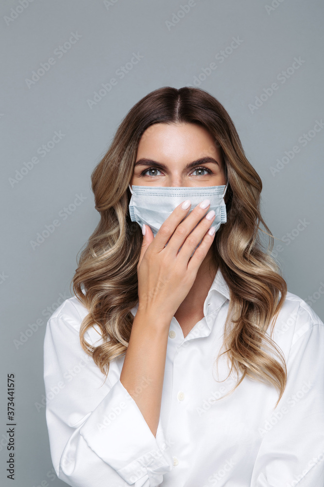 Confident young blonde woman with curly hair dressed in white shirt smiling, wearing protective mask against covid-19, hides a smile with her hand, looking at camera, isolated on grey background. 