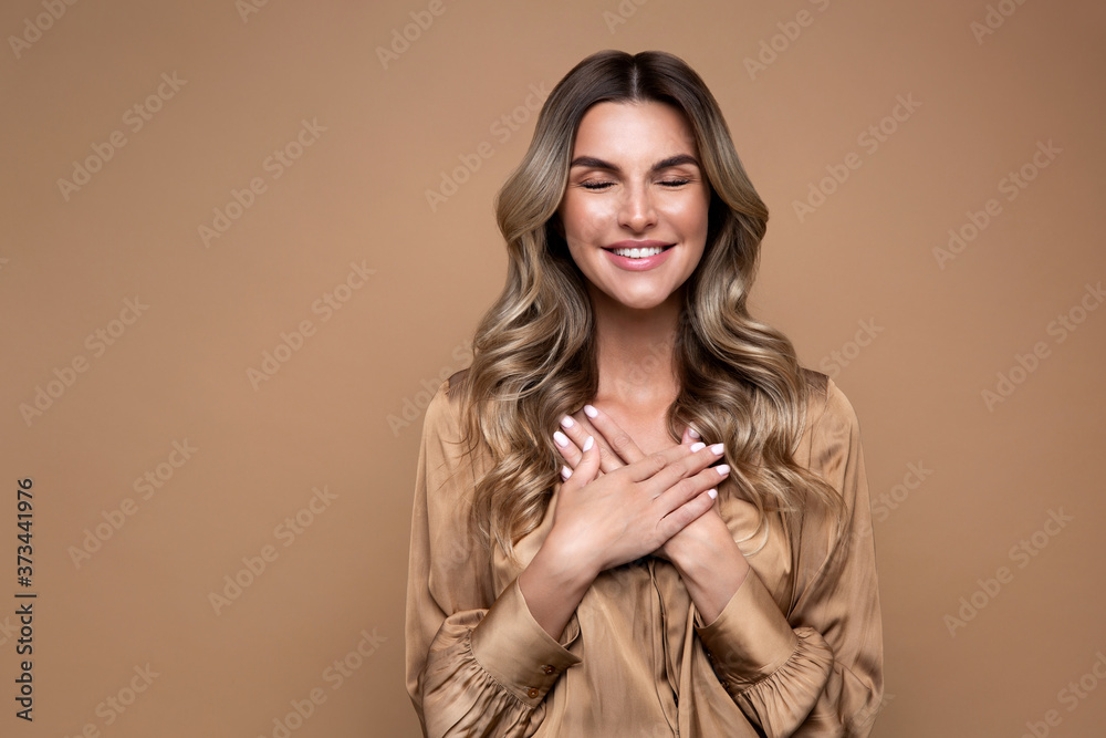 Happy smiling woman with curly hair and closed eyes keep hands at chest at heart, expresses sincere heartfelt appreciation, dressed in beige silk shirt, isolated on beige studio background. Happy smiling woman with curly hair and closed eyes keep hands at chest at heart, expresses sincere heartfelt appreciation, dressed in beige silk shirt, isolated on beige studio background.