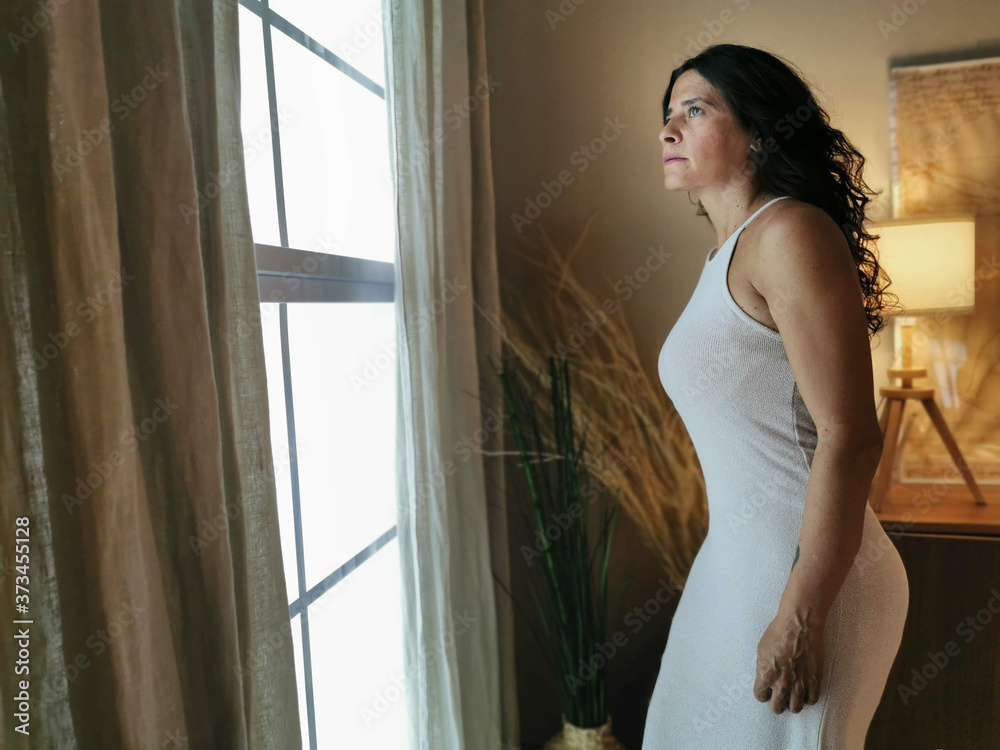 Woman looks out the window of her home opening the curtains in a home environment Woman looks out the window of her home opening the curtains in a home environment