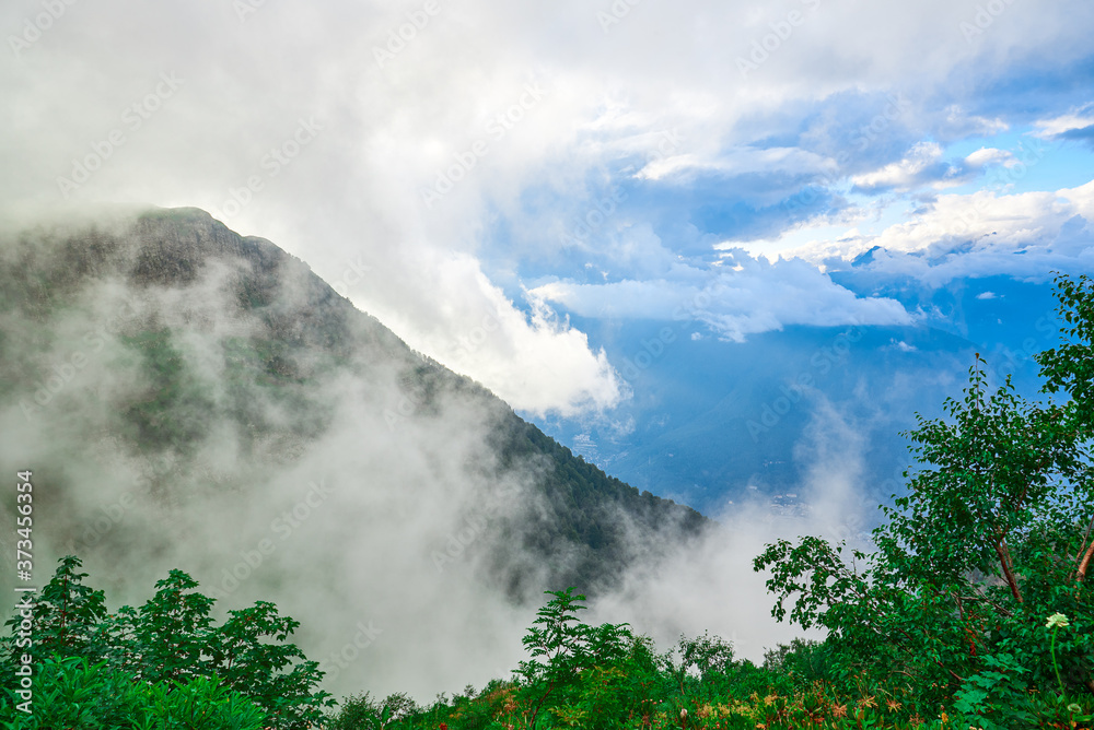 A high-altitude landscape with warm sunlight breaking through low clouds, fog creeping down the slope of a mountain covered with trees and a view of the valley below.