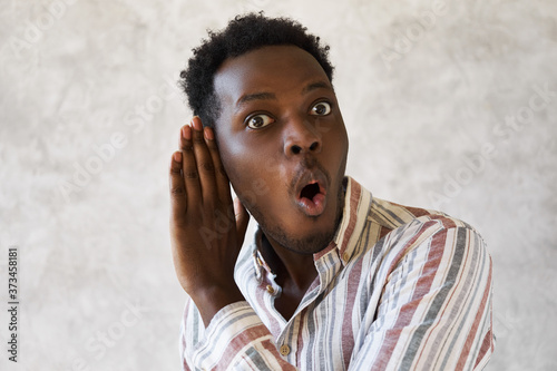Emotional curious young Afro American man with mouth gaped open expressing surprise and full disbelief holding hand at his ear overhearing secret private conversation, being totally shocked