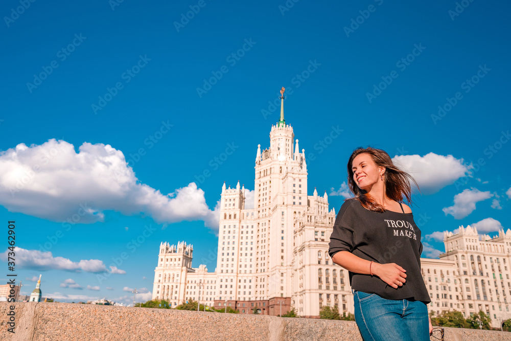 Obraz premium A young woman in a black jacket with an inscription walks along the embankment with a view of the Moscow river and the Stalin high-rise in Moscow