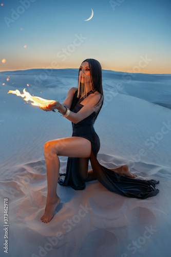 Fotografie A mysterious woman sits on the sand and holds a fire in hands