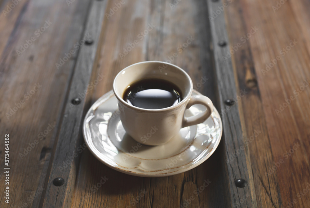 The white cup with black coffee and saucer are on a stylish wooden table. A coffee cup lit with daylight. Copy space.