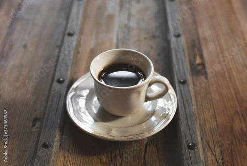 The white cup with black coffee and saucer are on a stylish wooden table. A coffee cup lit with daylight. Copy space.