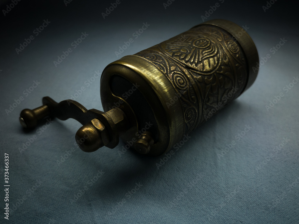 A small brass hand-held coffee grinder embossed on textiles against darkness. Traditional Turkish hand mill.