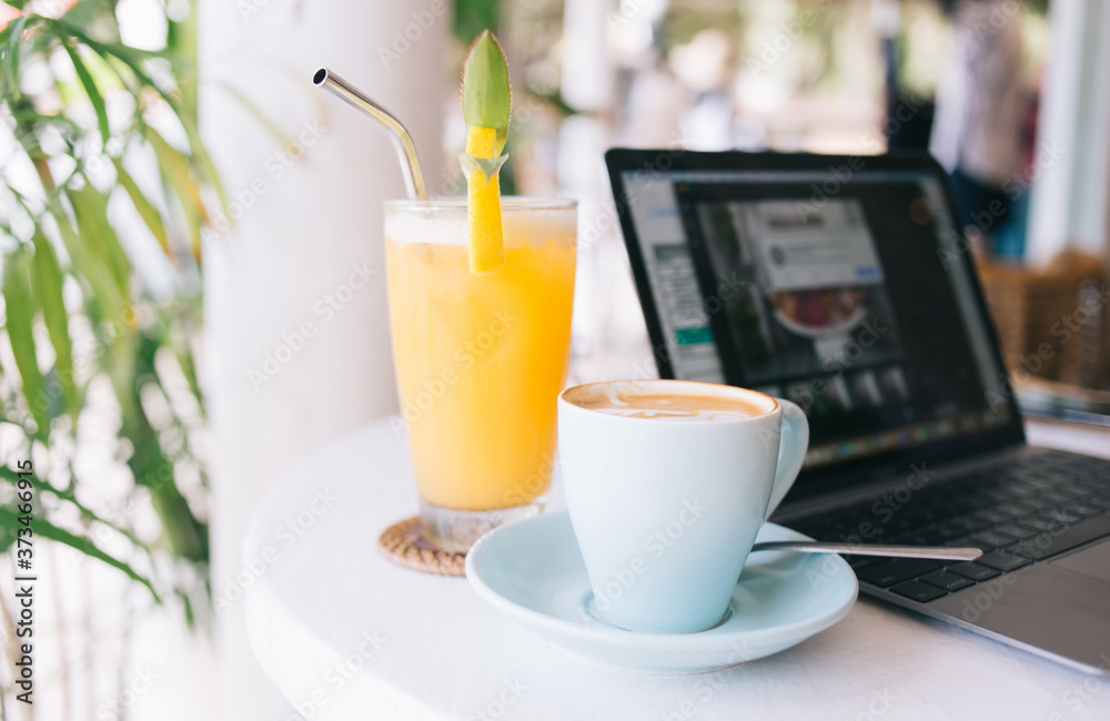 Cropped image of mug with coffee and orange juice for french breakfast near laptop computer on table, close up picture of fresh organic smoothie with straw in glass and caffeine cappuccino cup Cropped image of mug with coffee and orange juice for french breakfast near laptop computer on table, close up picture of fresh organic smoothie with straw in glass and caffeine cappuccino cup