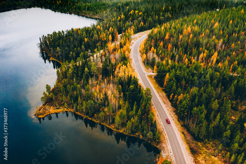 Aerial view of rural road with red car in yellow and orange autumn forest with blue lake
