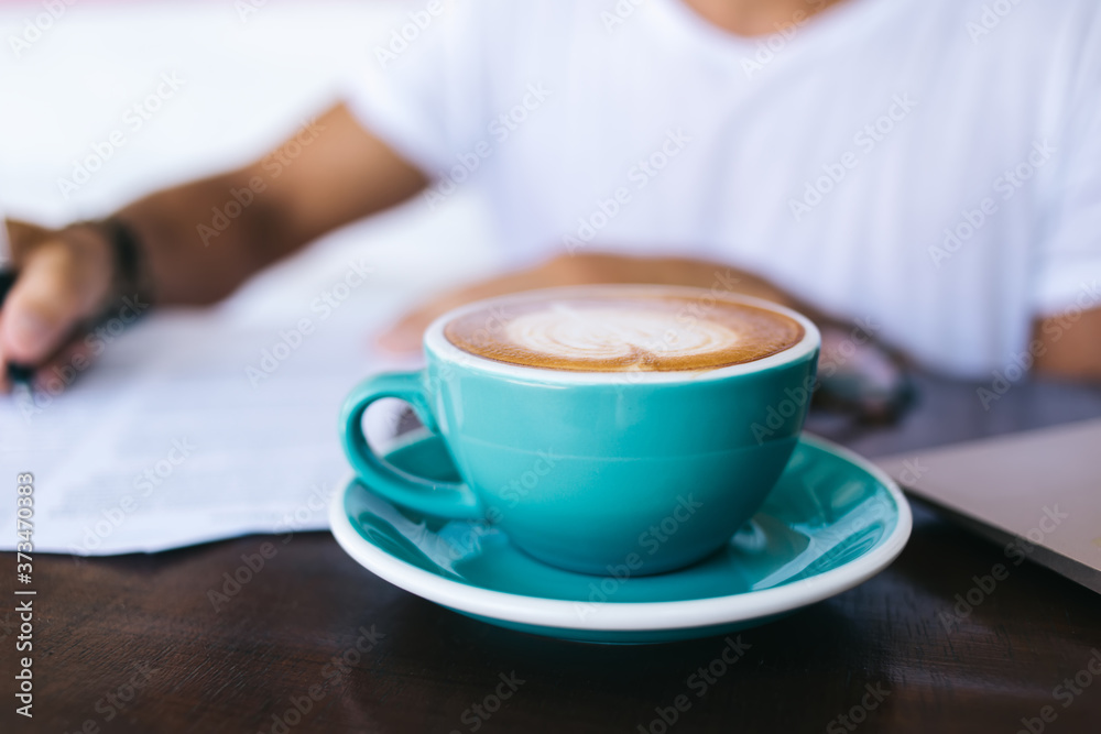 Selective focus on blue cup with caffeine beverage standing on working place with milky foram with coffee art on top, cropped image of hot coffee standing on blurred background with writing hand Selective focus on blue cup with caffeine beverage standing on working place with milky foram with coffee art on top, cropped image of hot coffee standing on blurred background with writing hand