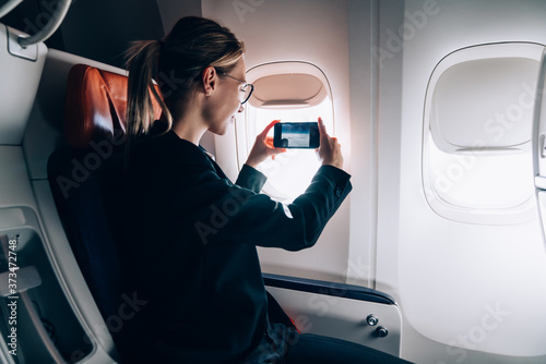 Pleased adult woman taking shot with smartphone through window in airplane