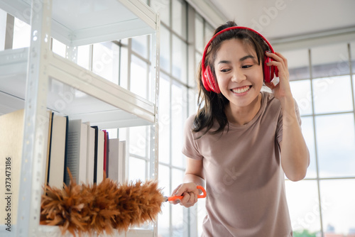 Young asian woman with headphones using a feather duster to clean wood book shelves in home .