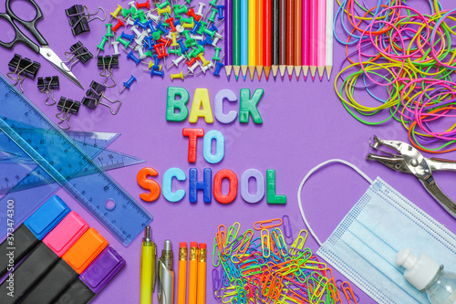School stationery supplies with medical mask and hand sanitizer on purple background. Back to school and new normal concept. COVID-19 prevention.  Top view with copy space