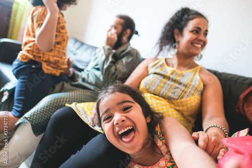 Happy female kid taking selfie with mobile phone camera at home with brother, mother and father in background - Cheerful indian family having fun sitting on couch at home - Soft focus on child face